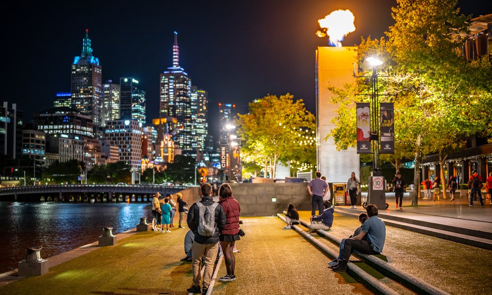 People sitting by the river at night watching flames erupt from towers with the city skyline in the background.