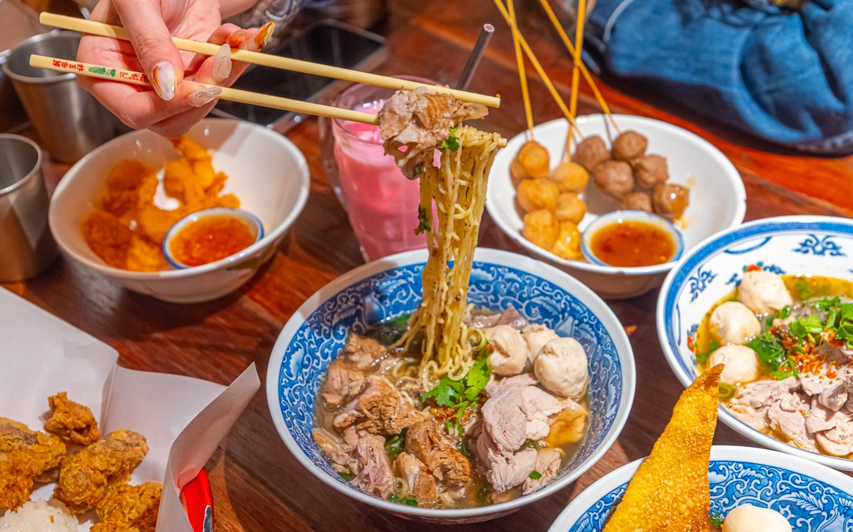 A table set with Thai dishes and someone lifting noodles from a bowl of soup.