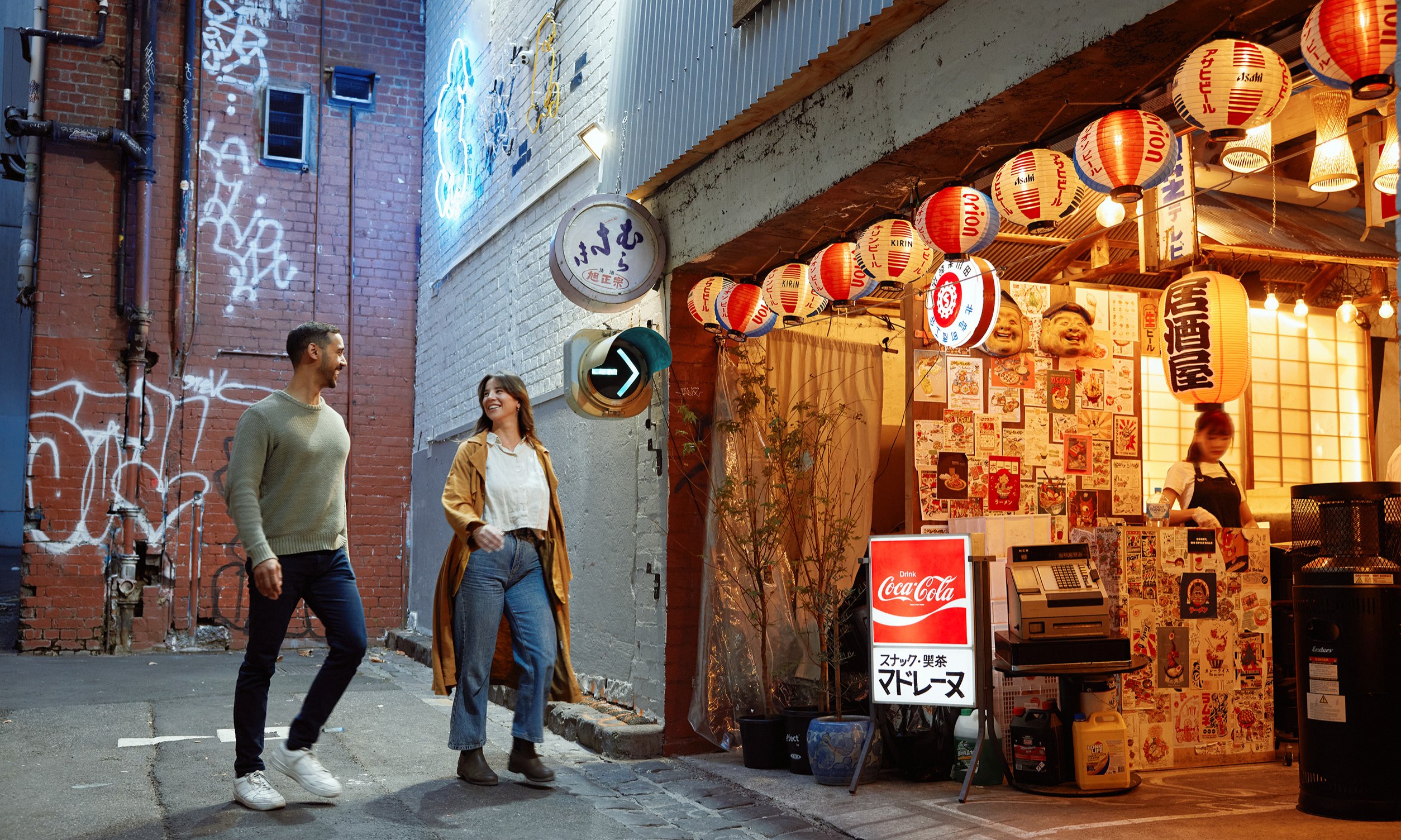 Two friends are walking into a small restaurant in a laneway. There are glowing lanterns hanging outside.