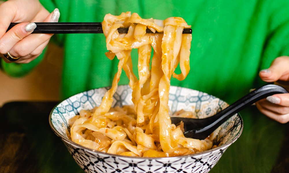 Person wearing a green jumper and picking up a bunch of noodles with chopsticks from a large noodle bowl.