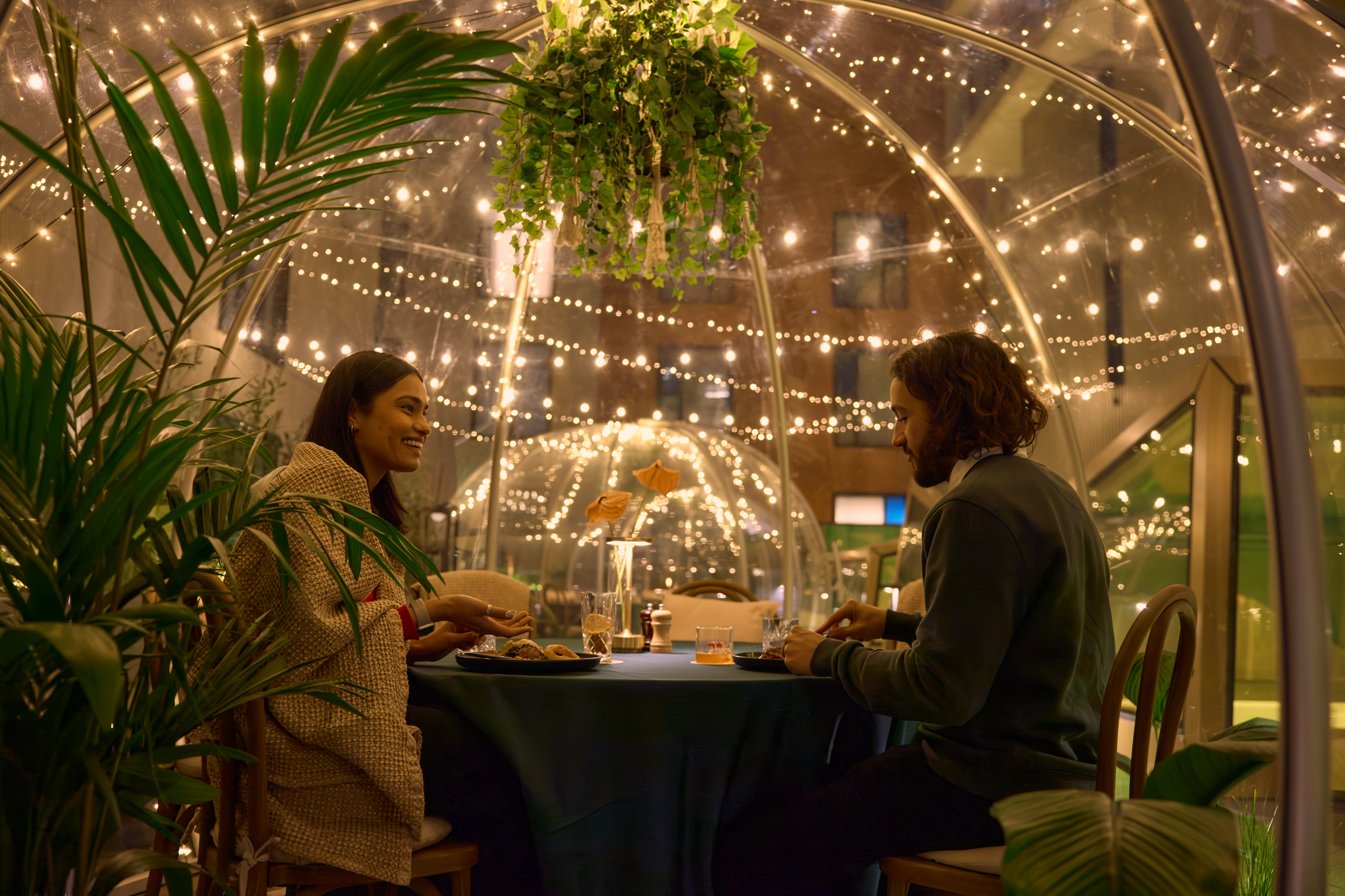 Two diners sit at a table under a dome decorated with fairy lights and plants