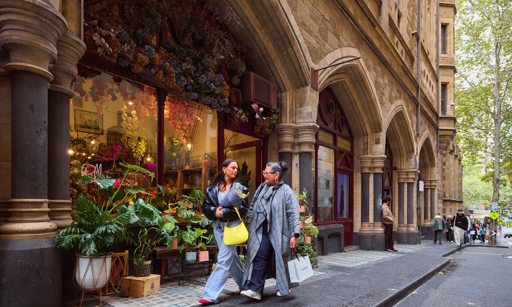 Two women walking past a florist with shopping bags and a bunch of flowers in their arms.