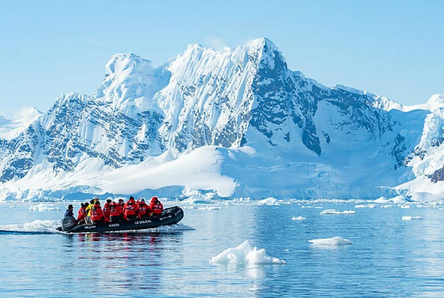 A motorised rubber dinghy travelling on calm sea past a glacier in Antarctica, carrying about a dozen people in red jackets.