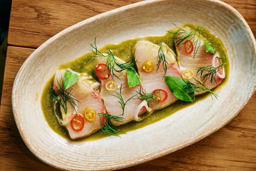 Four slices of Kingfish ceviche with sliced chillies, dill and a green sauce, on an oval platter.