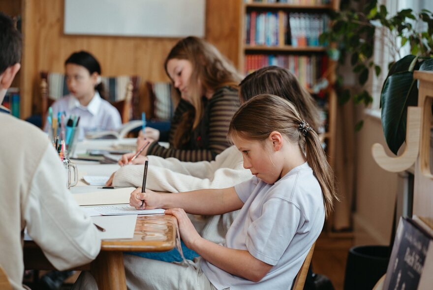 Kids seated around a large table writing and drawing during a creative writing workshop in a bright studio space, bookshelves in the background..
