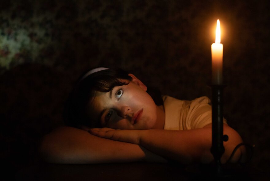 Person resting their head on their arms which are crossed flat on a table in front of them, a lit candle in the foreground.