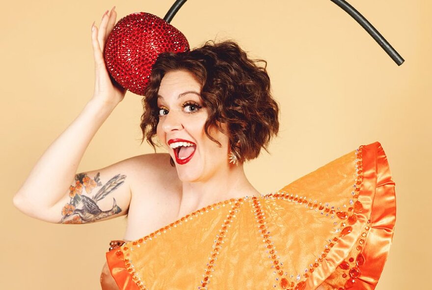 Burlesque performer hiding behind a large silk fan, holding a large sequinned cherry to her head with a tattooed arm.