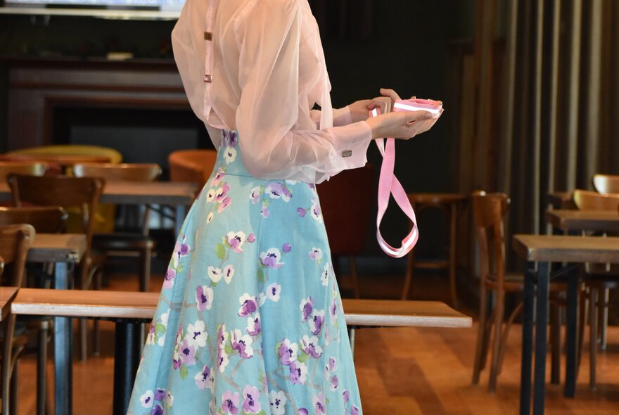 A woman's body, wearing a blouse and a blue floral skirt, a pink ribbon in her hands