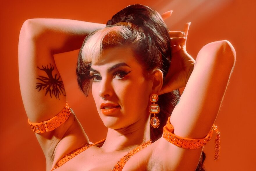 A burlesque performer dressed in orange against an orange background, her hands gathering up her hair behind her head. 