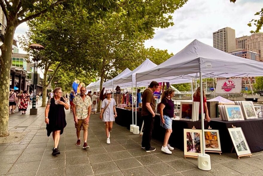 People walking past market stalls under white gazebos at an outdoor market, leafy trees on the left.