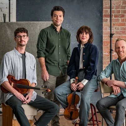 Four musicians of the Australian String Quartet posing for a photo, two of them holding their instruments, an  exposed brick wall behind them.