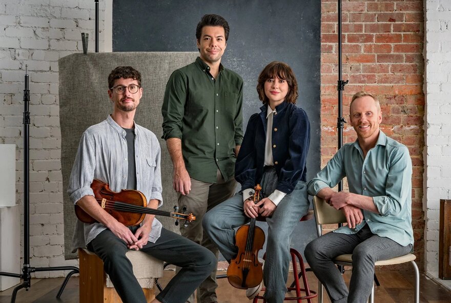 Four musicians of the Australian String Quartet posing for a photo, two of them holding their instruments, an  exposed brick wall behind them.