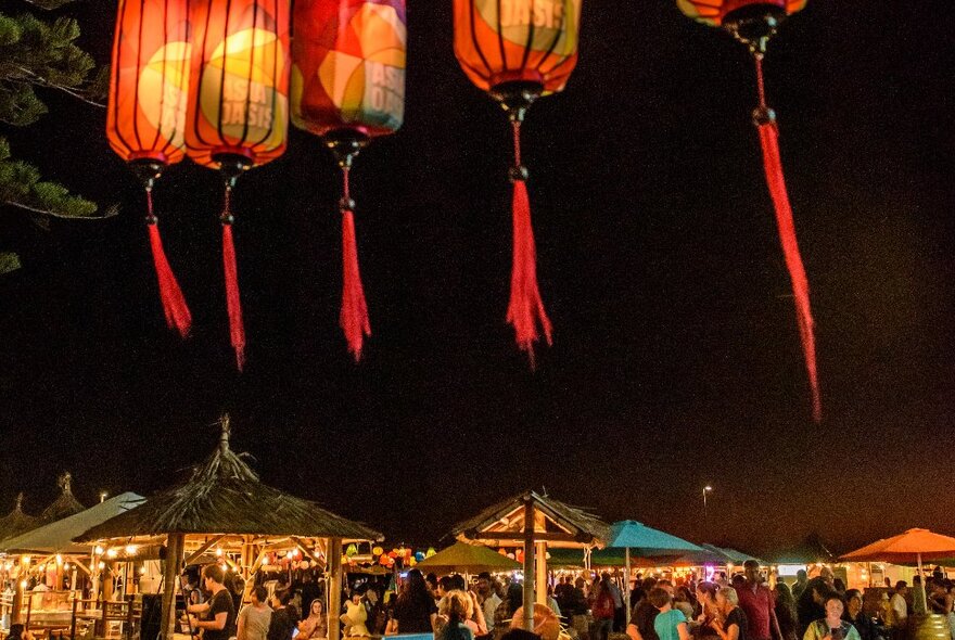 A collection of lit up night market stalls, with red lanterns hanging in the foreground.