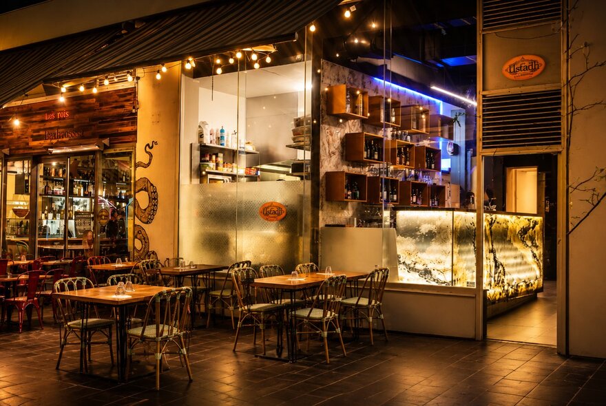 An outdoor restaurant dining area with wooden tables and chairs, inside is a brightly lit counter and shelves filled with bottles.