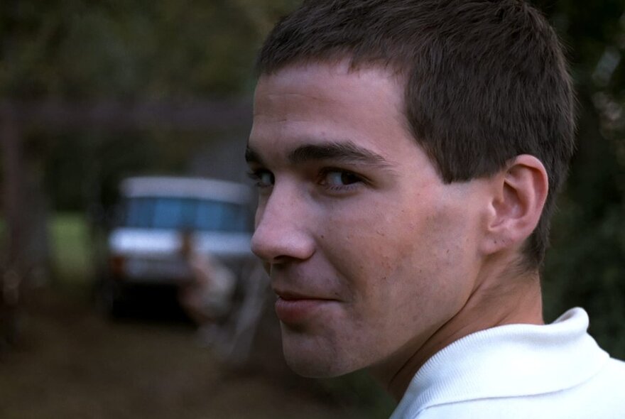 A still image from a film of a young man looking over his left shoulder and smirking. 