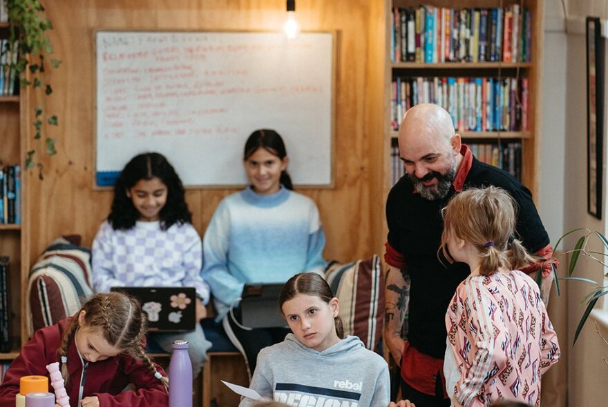 Primary school-aged children sitting around worktables, writing and interacting with each other and their laptops, an adult teacher nearby, bookshelves and a whiteboard on a rear wall.