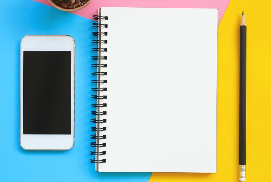 A mobile phone, an open, unlined notebook and a sharpened grey-lead pencil, on a blue, yellow and pink surface.