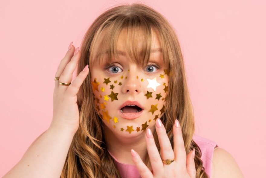 A person with long blonde hair and gold star stickers on their face, posing in front of a pink background with a surprised expression.
