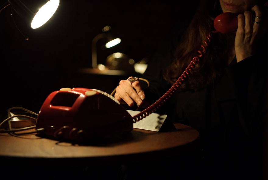 A seated person in shadows, holding up the earpiece of a red vintage phone, partly illuminated by a desk lamp, part of an interactive theatre installation at the Malthouse Theatre.
