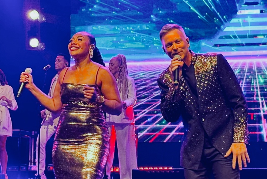 Singers Paulini and Tim Campbell on stage with back-up singers and a projection of bright neon lights behind them.