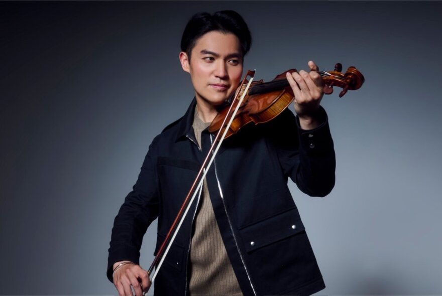 Classical violinist playing a bow across a Stradivarius, wearing a casual dark jacket.