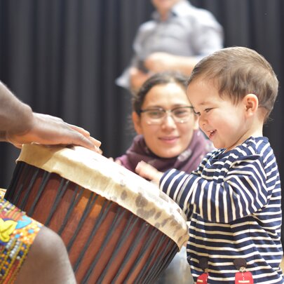 A standing toddler banging on an African drum while an adult woman looks on encouragingly.