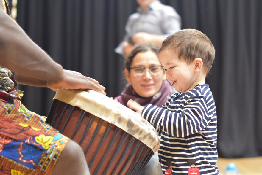 A standing toddler banging on an African drum while an adult woman looks on encouragingly.
