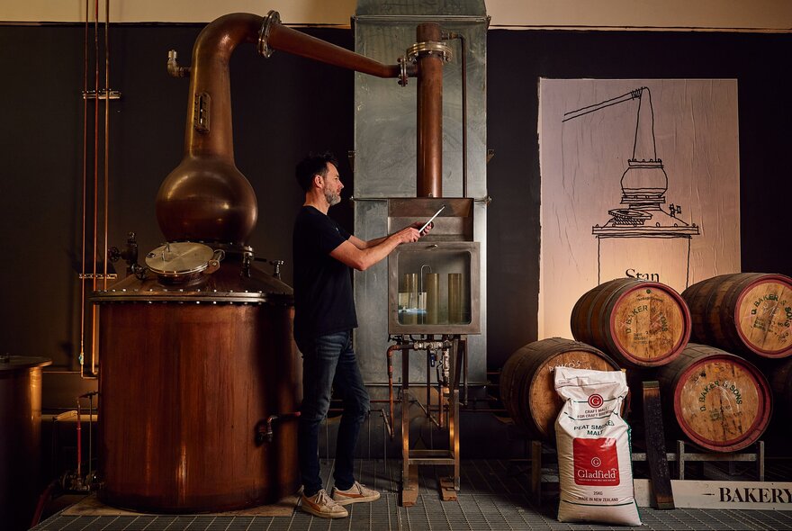 A man stands next to a large copper still and wooden barrels in a distillery.