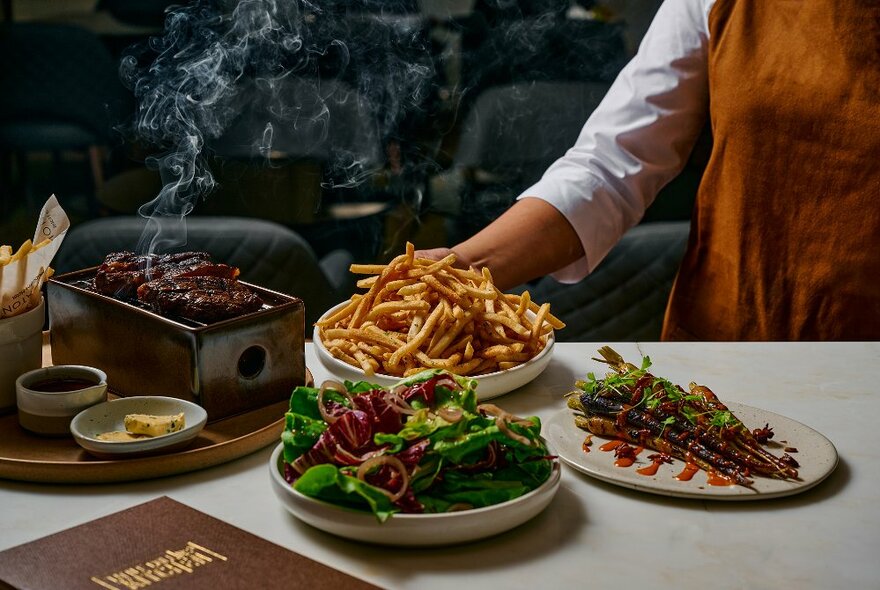 Server's hand placing a large bowl of chips on a table next to a steaming grill with meat and sides of salad and vegies.