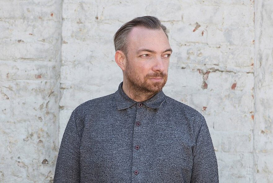 Man with short hair and a beard, wearing a grey shirt buttoned to the neck, looking sideways.