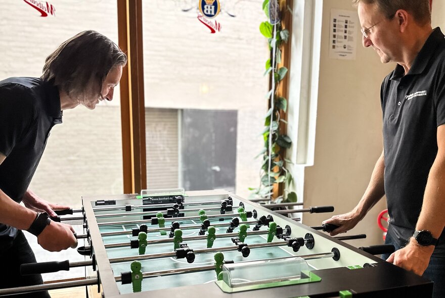 Two people playing Foosball in front of a window. 