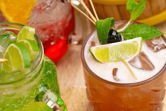 Close-up of two cocktails, one green with lime slices and the other brown with a lime wedge, mint, and a cherry.