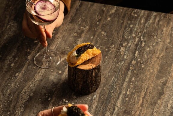 A caviar bite served on a rustic wooden block alongside a garnished cocktail.