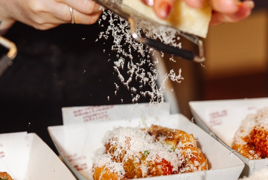 Hands grating cheese over a sausage in a roll. 