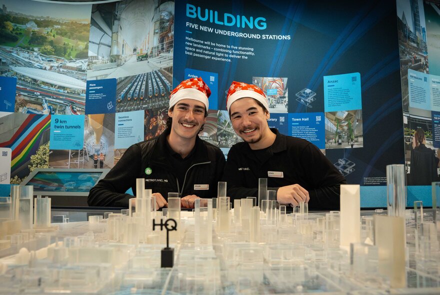 Two smiling men wearing Santa hats in front a table holding a glass model. 
