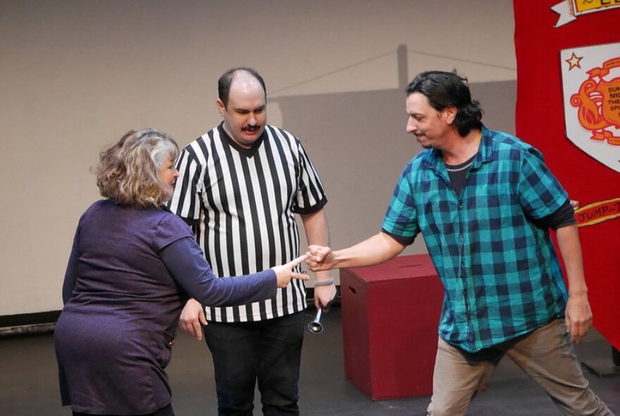 Three actors on stage performing an improvised theatre routine, one of theme dressed in a black and white striped top like a sports referee.