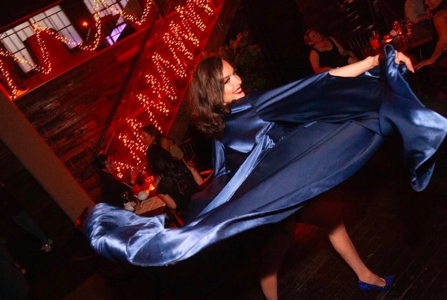 Burlesque dancer, Brook Bird, swirling a blue satin cape in a bar lit up by strings of fairy lights.