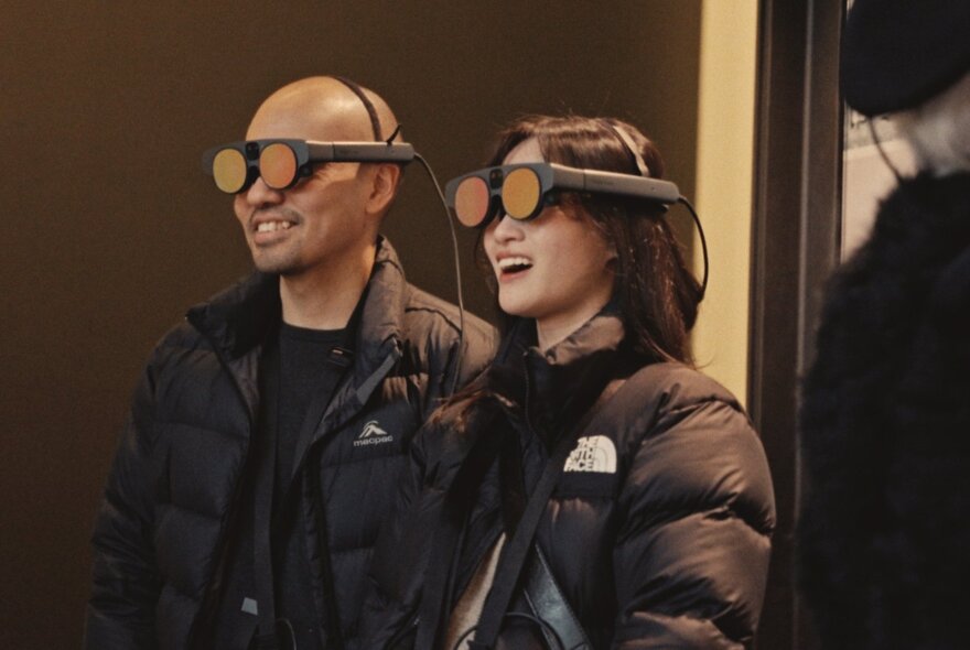 Two people wearing mixed reality headsets and black puffer jackets.