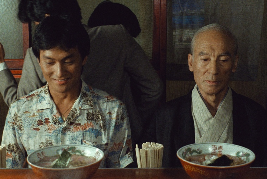 Two Japanese men, one younger, one older, seated at a table in a ramen restaurant in Japan, each seemingly unsatisfied with the bowl of ramen in front of him. 