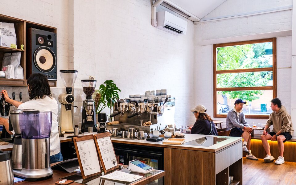 Inside a cafe. There is a barista making coffee and people sitting down drinking coffee.