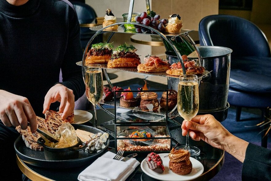 Hands holding wine glasses and bread at a restaurant table loaded with food in display cases plus crockery and napkins.