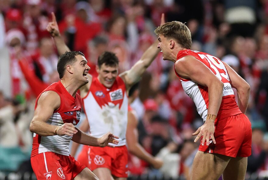 A group of Sydney Swans AFL football players high-fiving and celebrating in a huddle on the field with blurred fans in the stadium in the background.