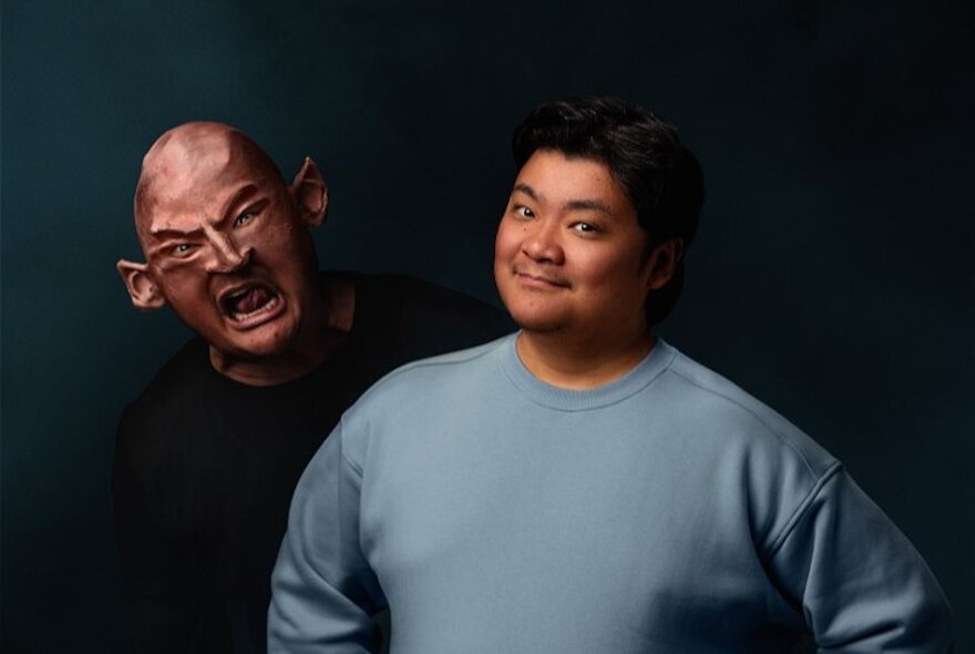 Performer Randy Adeva smiling, wearing a grey long-sleeved top, behind him is a person wearing a scary horror mask.
