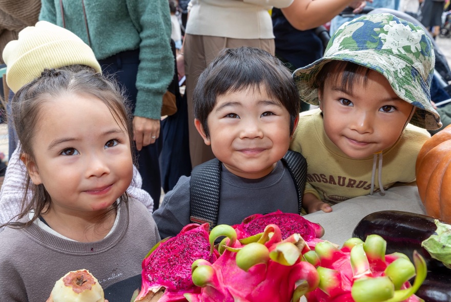 Three young children smiling in front of a table of dragon fruit at an outdoor festival.