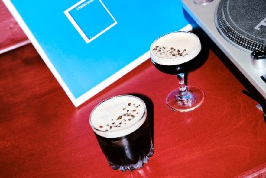 Two espresso martini-like cocktails in different glasses on a red table with a blue card. 