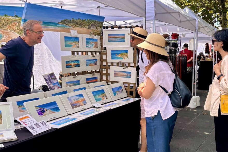 People browsing seaside landscape artworks at an outdoor market stall.