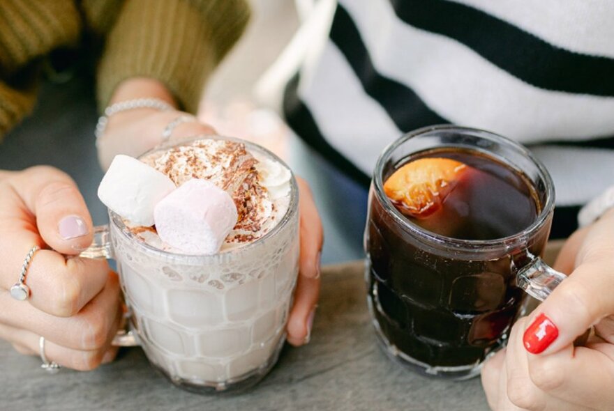 Two people holding glass mugs, one is filled with a milky drink with milo and marshmallows, the other is mulled wine with an orange wedge.