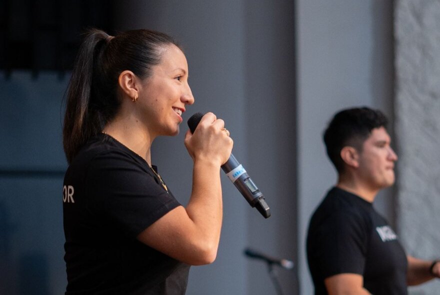 A woman speaking into a handheld microphone while wearing a short-sleeved black T-shirt, with a man in a matching black T-shirt standing in the background.