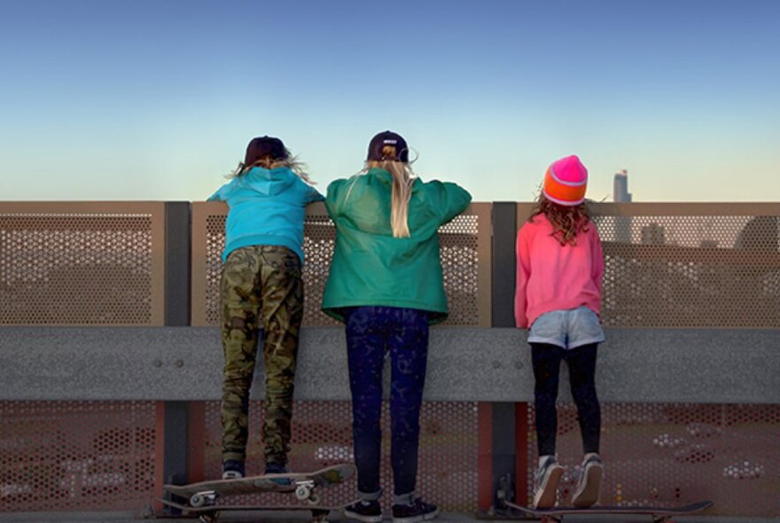 Three young, female Olympic skateboarding hopefuls, looking over the tall barrier of a bridge at the city behind.