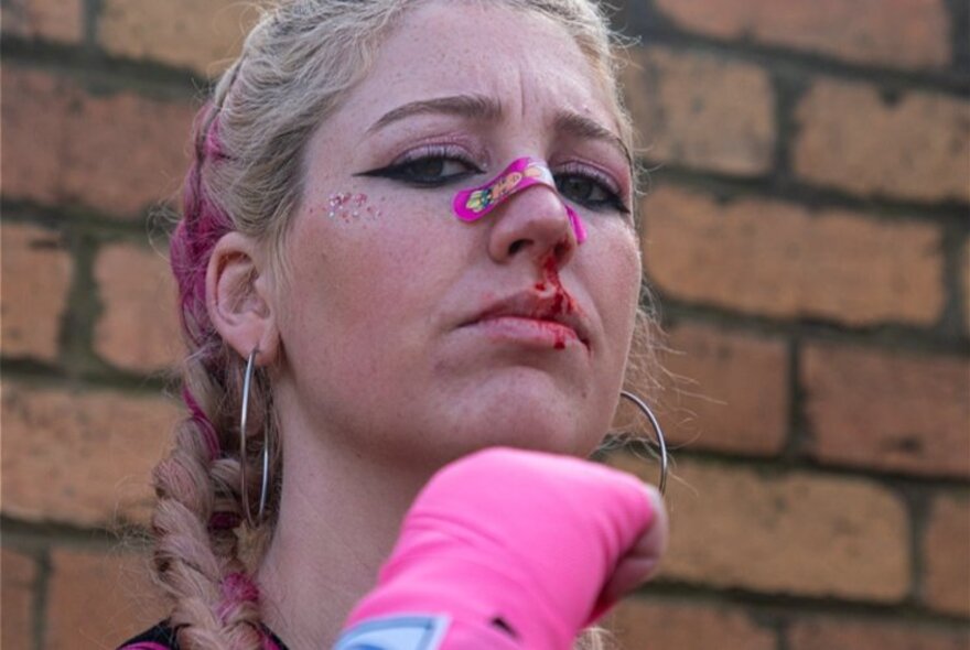 A serious woman looking like she's been in a fight with a bandaid on her nose and pink boxing gloves